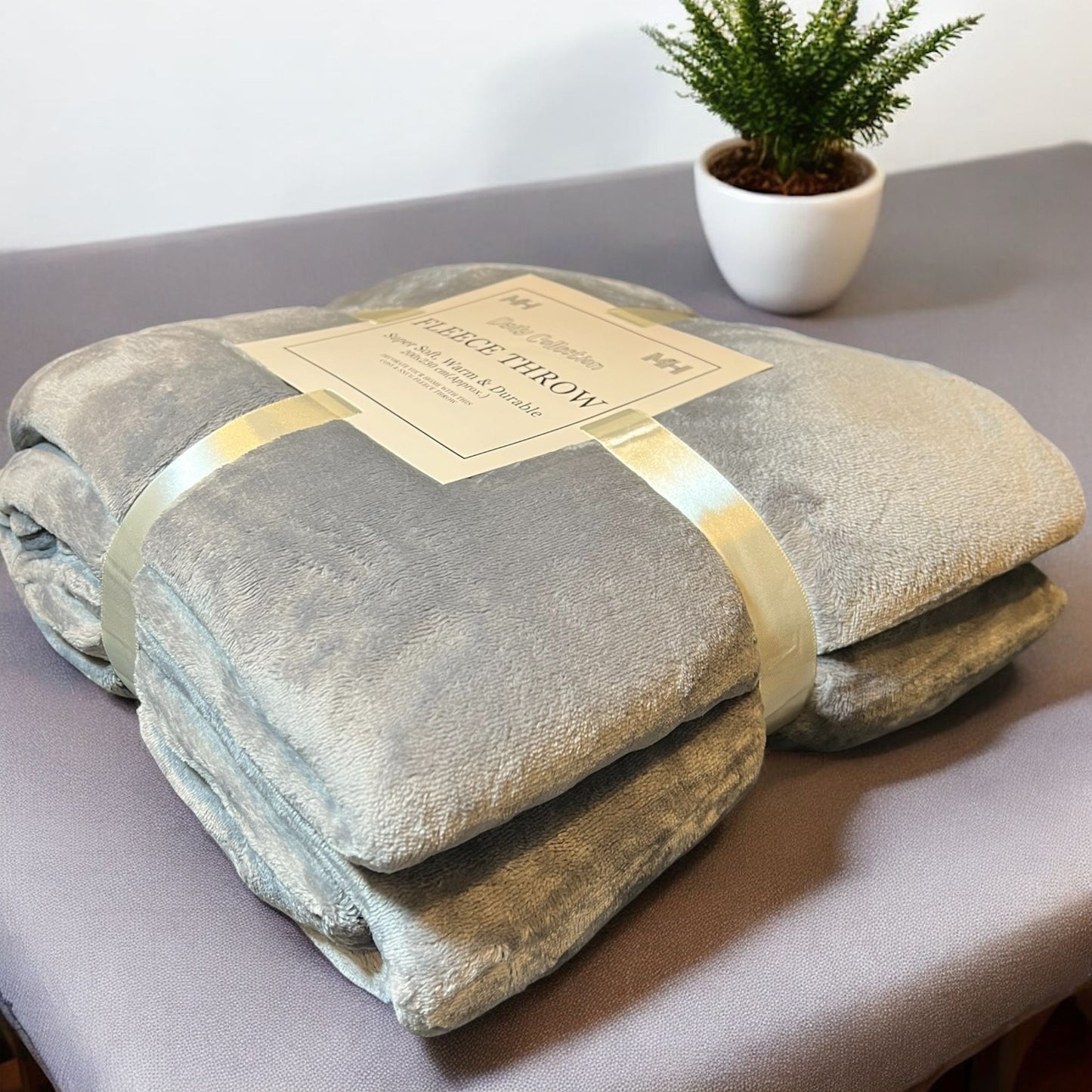 Folded plush throw blanket with a label on a gray surface, next to a small potted plant.