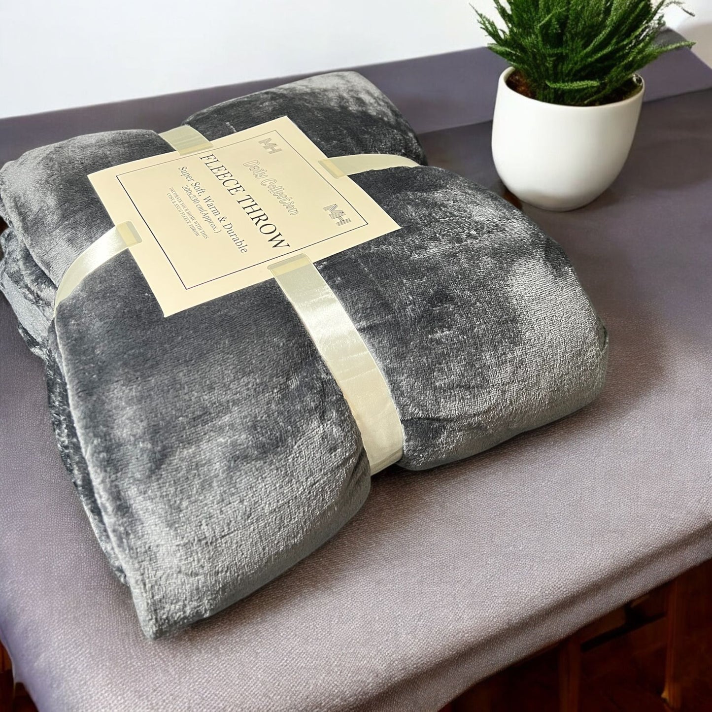 Gray fleece throw blanket with a label on a surface, next to a potted plant.