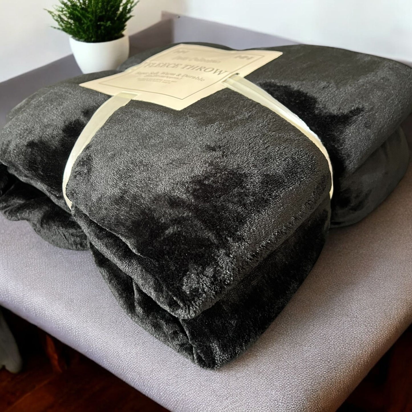 Folded dark gray blanket with a label on a light surface, with a plant in the background.