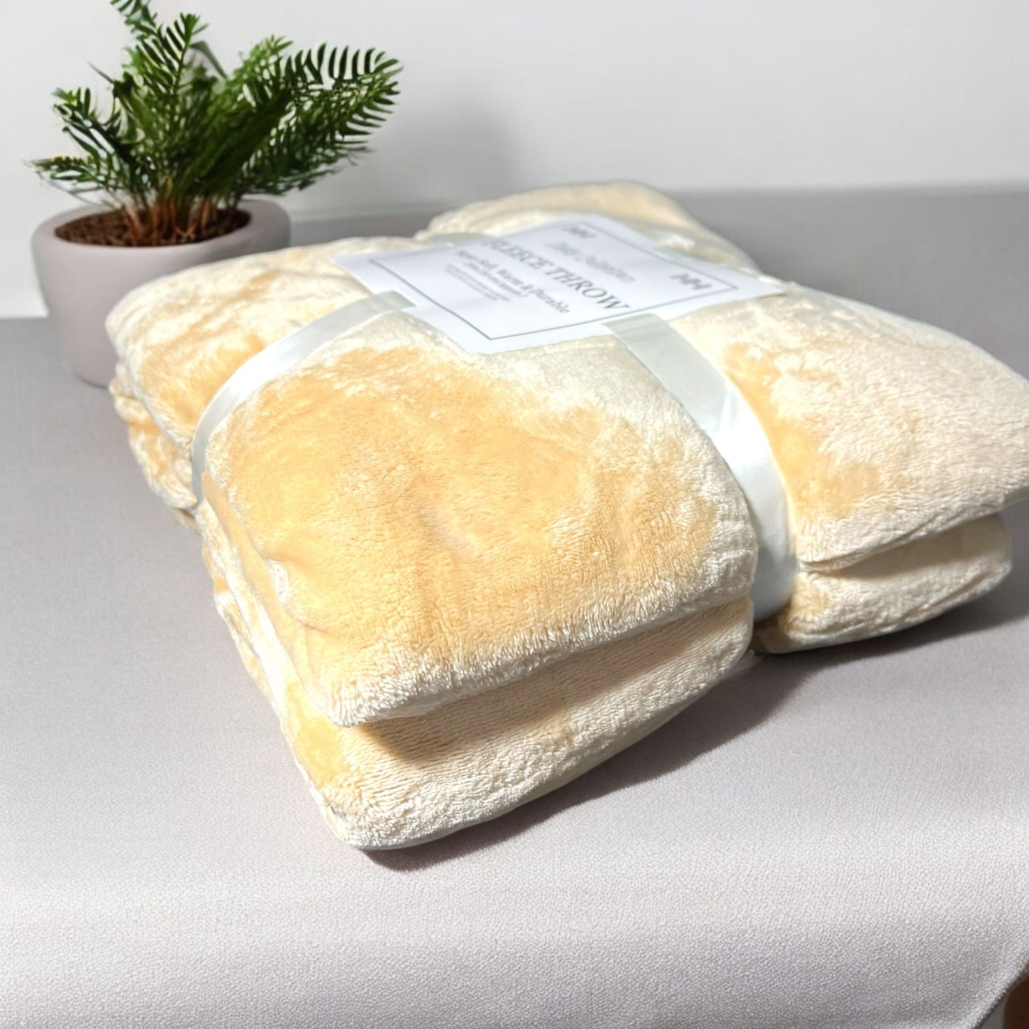 Folded beige blanket with a label on a light gray surface, next to a small potted plant.